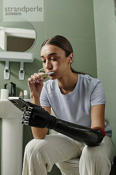 Young woman with black bionic arm brushing teeth and using smart phone in bathroom at home