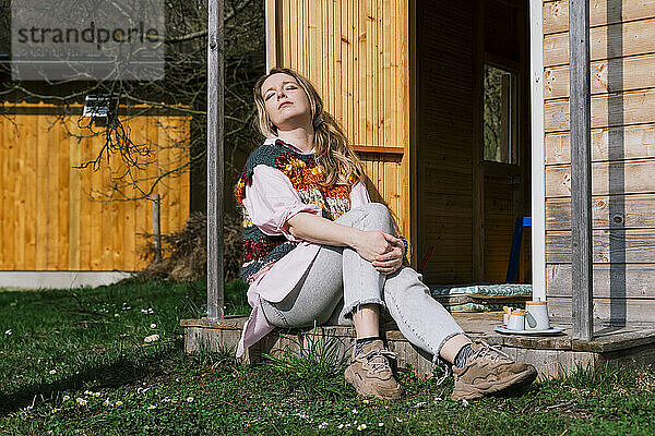 Woman with eyes closed resting on porch at back yard in sunlight