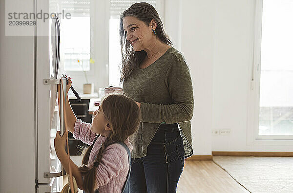 Granddaughter writing on refrigerator standing by grandmother at home