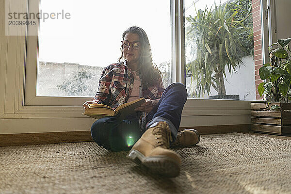 Mature woman reading book leaning on window at home