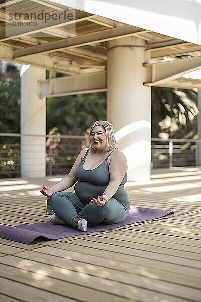 Smiling curvy young woman doing yoga on mat in park