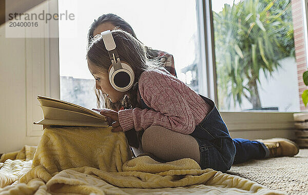 Girl listening to music through wireless headphones and reading book by grandmother at home