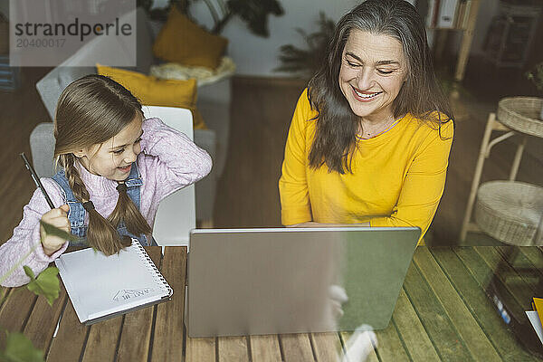 Grandmother and granddaughter using laptop together seen through window