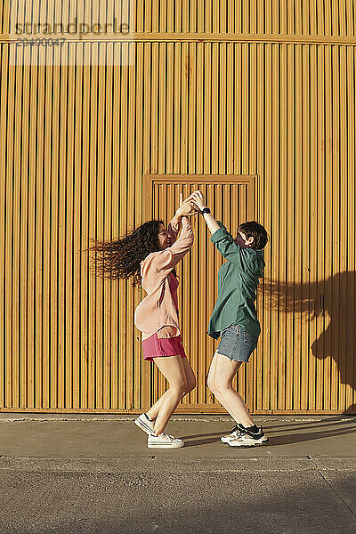 Happy gay couple dancing together with holding hands in front of brown building wall