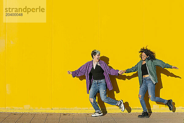 Cheerful lesbian couple running together in front of yellow wall