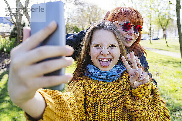 Cheerful girl showing peace sign and taking selfie through smart phone with mother at park