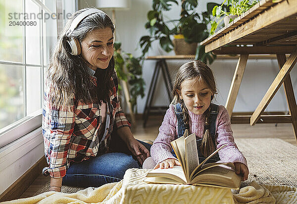 Grandmother listening to music sitting by granddaughter reading book at home