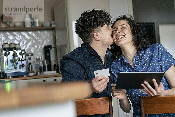 Man kissing girlfriend holding tablet PC in kitchen at home