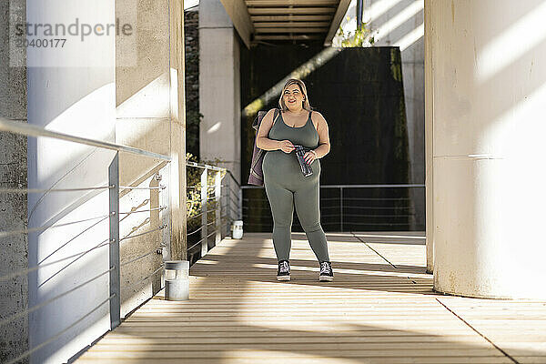 Smiling curvy woman standing with water bottle near railing