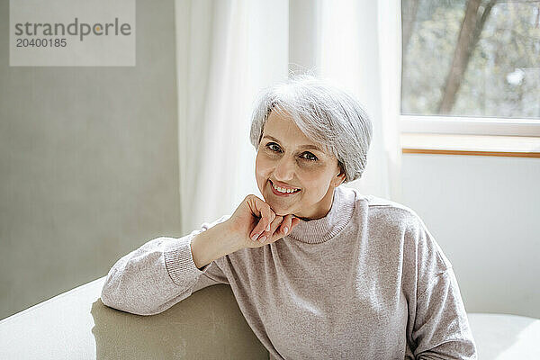 Smiling mature woman with hand on chin sitting on couch at home