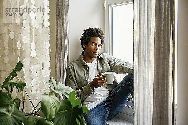 Man with coffee cup sitting on window sill at home