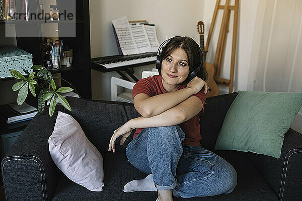 Thoughtful young woman with wireless headphones sitting on sofa in living room at home