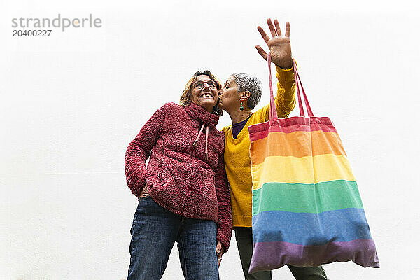 Woman holding multi colored bag and kissing girlfriend near wall