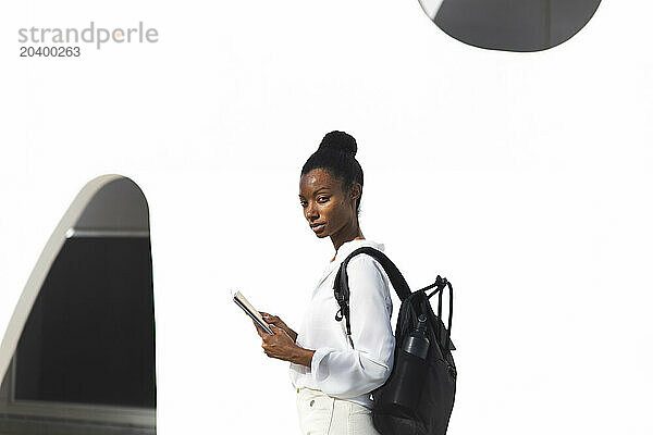 Confident mature woman with backpack and book in front of white building