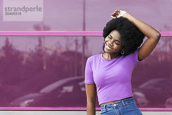 Happy young Afro woman with hand raised standing in front of magenta window