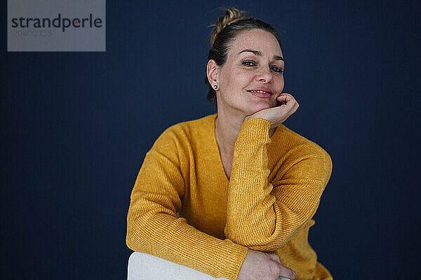 Smiling beautiful woman sitting with hand on chin against colored background