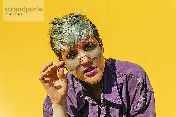 Woman with dyed hair holding sunglasses in front of yellow wall