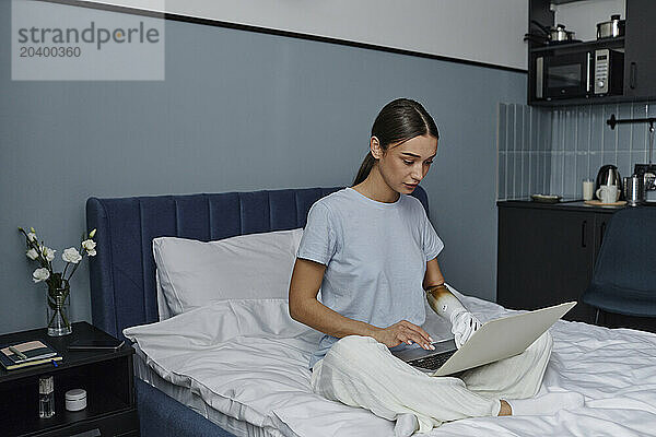 Woman with arm prosthesis using laptop sitting on bed in bedroom