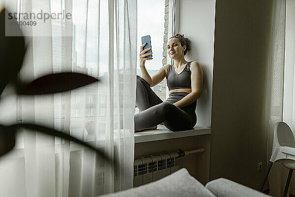 Woman using smart phone sitting on window sill at home