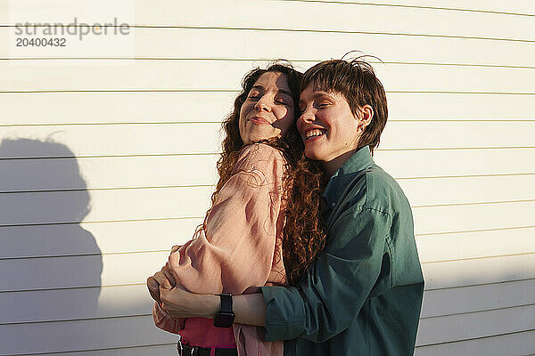 Happy lesbians embracing with eyes closed in front of white wall