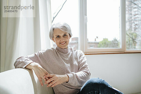 Smiling mature woman sitting on couch by window at home