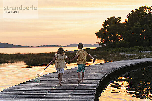 Brother and sister with fishing net running on pier over lake at sunrise