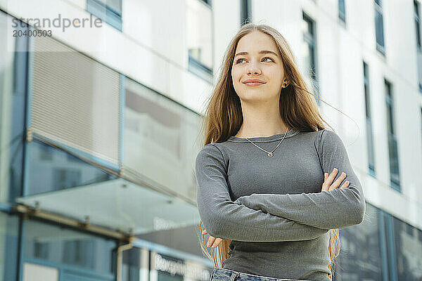 Smiling teenage girl with arms crossed looking away in front of building