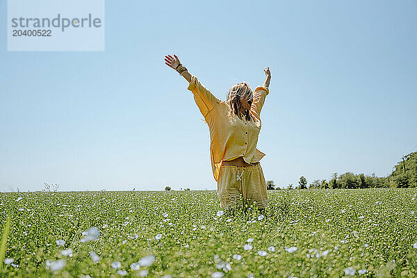 Happy woman wearing yellow linen clothing standing with arms raised in blooming field on sunny day