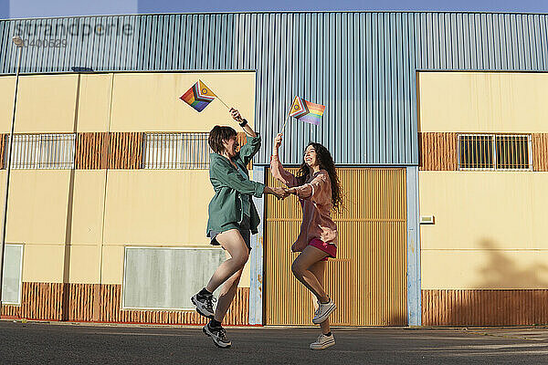 Happy lesbian couple dancing and raising LGBTQIA pride flag in front of building