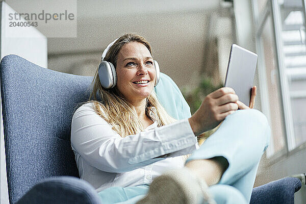 Happy businesswoman listening to music sitting with tablet PC at office