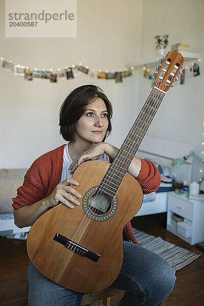 Thoughtful young beautiful woman with guitar sitting in bedroom at home