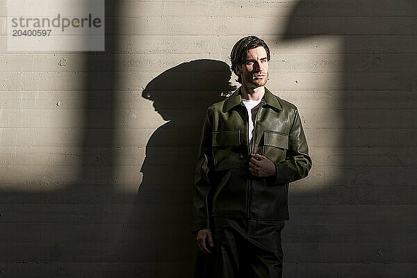 Fashionable young handsome man wearing green leather jacket leaning on wall