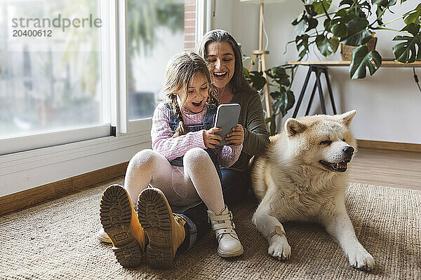 Happy grandmother and granddaughter using smart phone sitting by dog at home