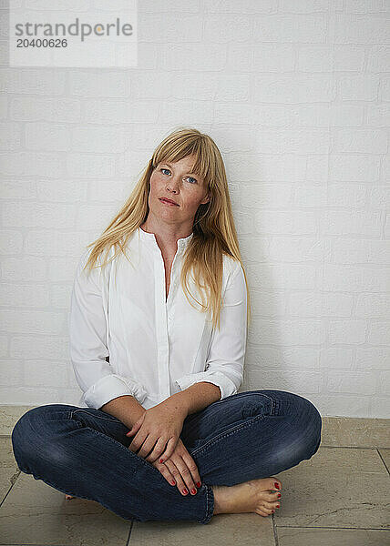 Mature woman sitting on floor with legs crossed near white wall