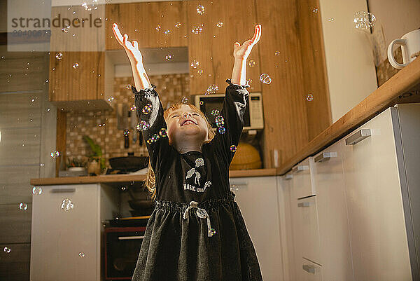 Happy girl smiling and playing with soap bubbles in kitchen at home