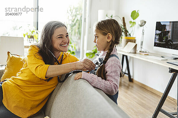 Happy grandmother listening to granddaughter's heartbeat through stethoscope at home