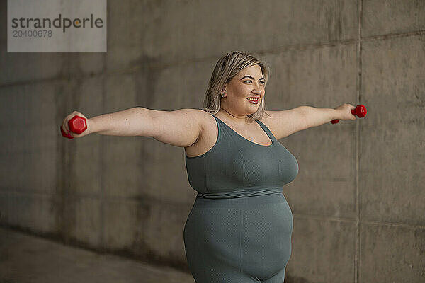 Smiling young curvy woman exercising with dumbbells near wall