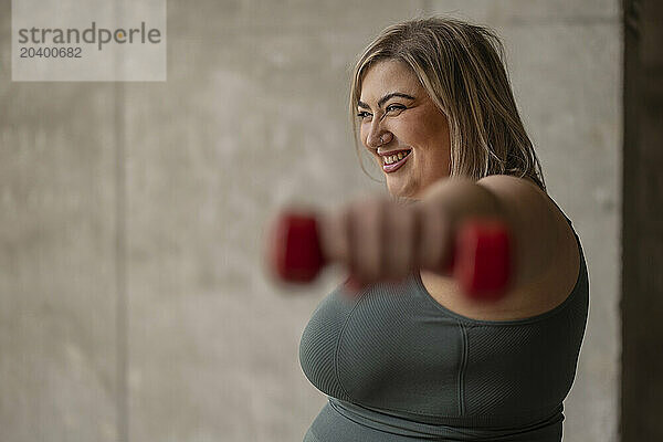 Happy curvy woman exercising with dumbbell near wall