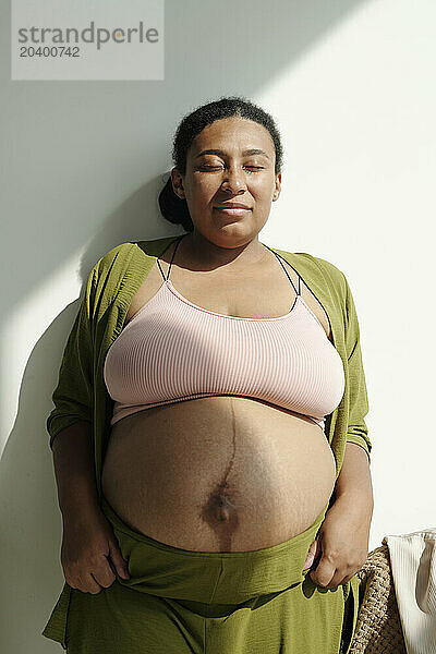 Plus size pregnant woman with eyes closed leaning on wall at home