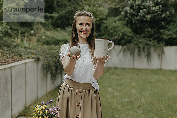 Smiling woman holding clay and ceramic cup standing at back yard