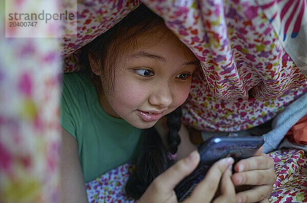 Curious girl using smart phone under blanket at home