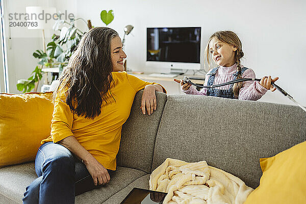 Smiling woman sitting on sofa looking at granddaughter playing with stethoscope in living room