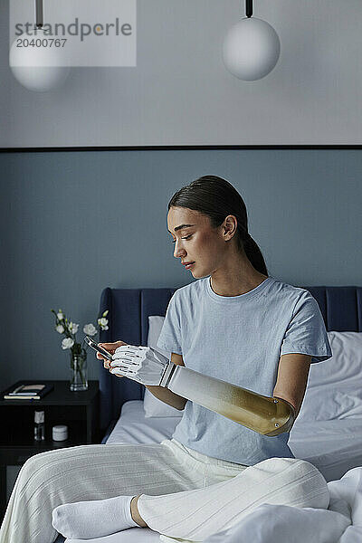 Woman with prosthetic arm using smart phone sitting on bed in bedroom