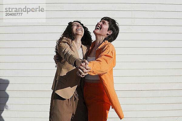 Happy lesbian couple holding hands dancing together in front of white wall