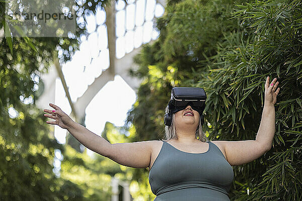 Smiling curvy woman wearing virtual reality simulator and gesturing near plants