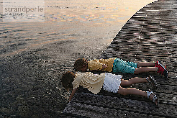 Brother and sister lying on pier fishing in lake at sunrise