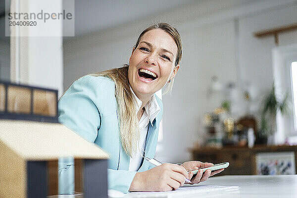 Happy mature architect sitting with smart phone near model house at desk in office