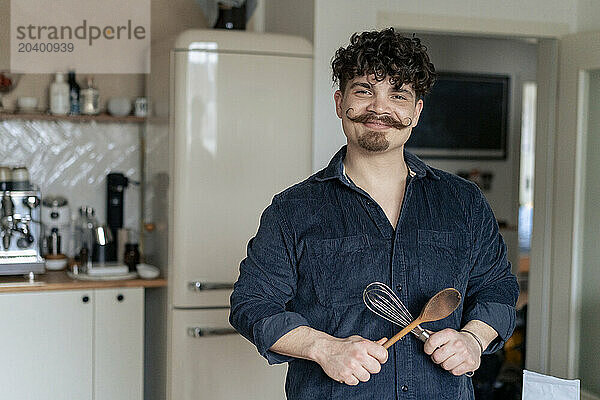 Smiling man holding wooden spoon and wire whisk in kitchen at home