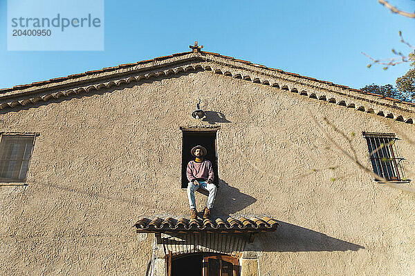 Man sitting on window of house under blue sky