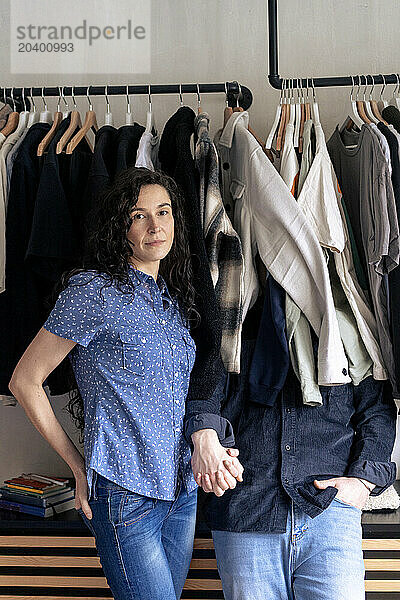 Woman holding hand of man hiding behind clothes at home